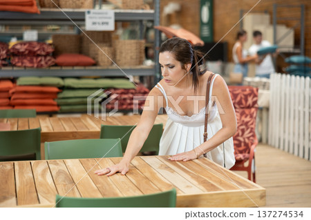 Pretty brunette girl in white dress chooses wooden table for country house in garden store Pretty brunette girl in white dress chooses wooden table for country house in garden store 137274534