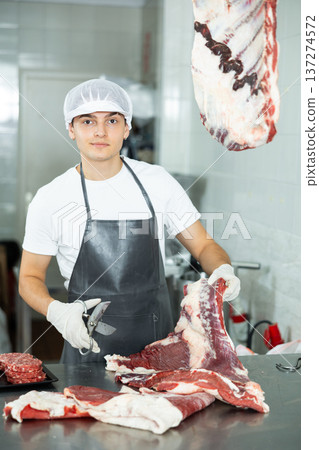 Guy employee of butcher shop cuts meat carcass into small pieces of meat for retail sale in store 137274572