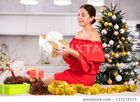 Emotional young female in red dress examining packed gift box with bow near decorated Christmas tree at home Emotional young female in red dress examining packed gift box with bow near decorated Christmas tree at home 137274573
