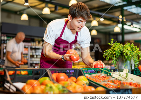 Skilled young seller arranging ripe red tomatoes on vegetable counter Skilled young seller arranging ripe red tomatoes on vegetable counter 137274577