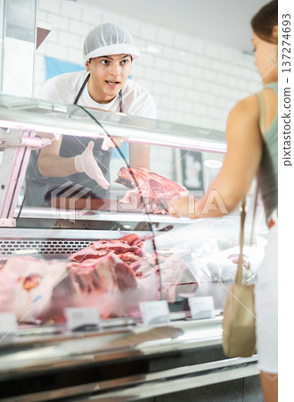 Positive young salesman demonstrating piece of meat to purchaser in butcher shop Positive young salesman demonstrating piece of meat to purchaser in butcher shop 137274693