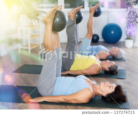 Old woman practicing pilates with softball between legs in light fitness room 137274723