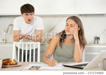 Young girl sitting at the table while her boyfriend is quarreling with her standing behind Young girl sitting at the table while her boyfriend is quarreling with her standing behind 137274860
