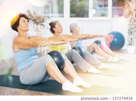 Old woman practicing pilates sitting with softball between knees in light fitness room Old woman practicing pilates sitting with softball between knees in light fitness room 137274916