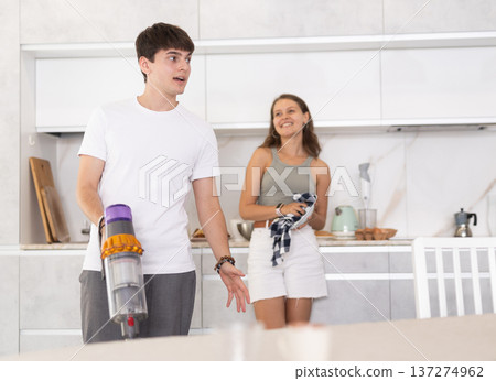 Guy vacuum-cleaning in the kitchen while communicating with his girlfriend drying dishes with towel Guy vacuum-cleaning in the kitchen while communicating with his girlfriend drying dishes with towel 137274962