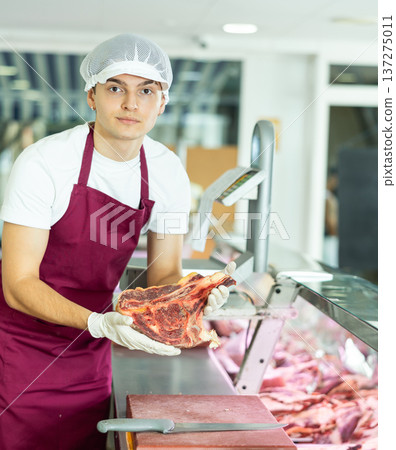 Guy employee of butcher shop cuts meat carcass into small pieces of meat for retail sale in store Guy employee of butcher shop cuts meat carcass into small pieces of meat for retail sale in store 137275011