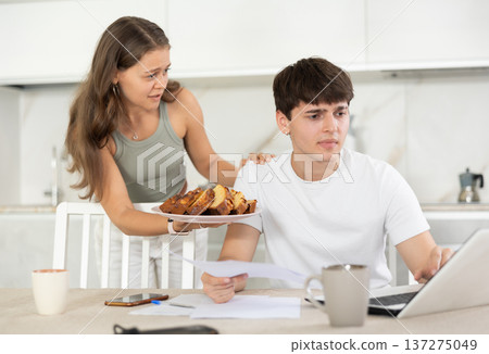 Busy guy working on notebook in the kitchen and a girl offering him pie Busy guy working on notebook in the kitchen and a girl offering him pie 137275049