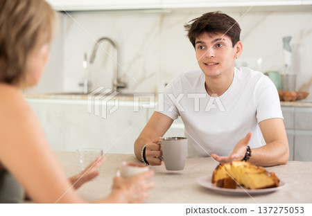 Adult woman and young guy drink tea and talk in kitchen Adult woman and young guy drink tea and talk in kitchen 137275053