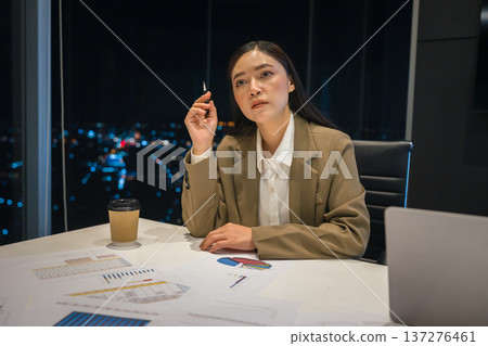 Tired businesswoman thinking and analyzing business reports late at night in office with city lights background Tired businesswoman thinking and analyzing business reports late at night in office with city lights background 137276461