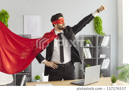 Funny confident businessman wearing red superhero cape and mask posing with raised fist in office. 137276537