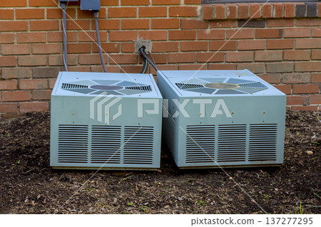 Two air conditioning units are installed against brick wall in yard area 137277295