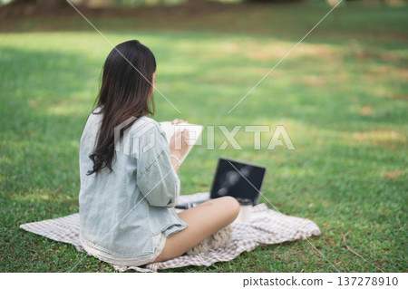 Young Woman Sketching in a Green Park While Sitting on a Blanket with Laptop and Art Supplies Surrounded by Nature and Sunshine 137278910