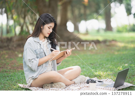Young woman writing in notebook while sitting on grass in park with laptop and coffee cup, enjoying peaceful outdoor workspace in nature 137278911