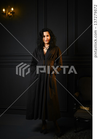 Woman stands in a dark room with hands in pockets wearing a long black dress during evening hours Woman stands in a dark room with hands in pockets wearing a long black dress during evening hours 137279872