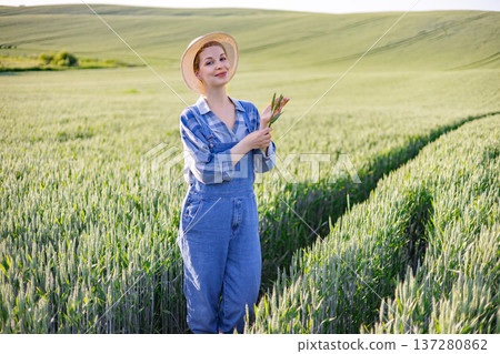 A young woman wearing a straw hat and denim overalls stands in a lush green wheat field, holding a few stalks of wheat A young woman wearing a straw hat and denim overalls stands in a lush green wheat field, holding a few stalks of wheat 137280862