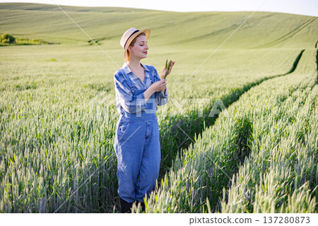A young woman wearing overalls and a straw hat stands in a vast green wheat field, examining a stalk of wheat A young woman wearing overalls and a straw hat stands in a vast green wheat field, examining a stalk of wheat 137280873