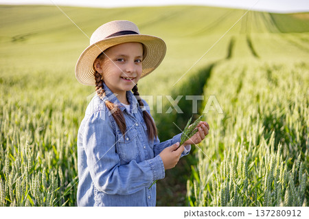 A young girl wearing a straw hat and denim shirt stands in a lush green wheat field, holding a stalk of wheat A young girl wearing a straw hat and denim shirt stands in a lush green wheat field, holding a stalk of wheat 137280912