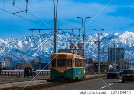 一輛電車駛過富山市的久禮羽橋，背景是白雪皚皚的立山山脈。 137284444