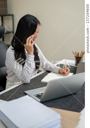 Young asian businesswoman office worker talking on phone call while using a laptop at company table. Young asian businesswoman office worker talking on phone call while using a laptop at company table. 137284600