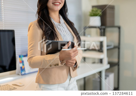 Close up of smiling asian businesswoman or office worker holding tablet standing at computer table. 137284611