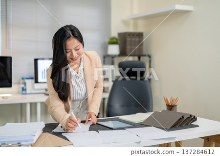 Smiling asian businesswoman or office worker holding pen over document standing leaning over table. Smiling asian businesswoman or office worker holding pen over document standing leaning over table. 137284612
