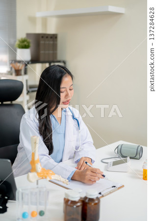 Young asian woman doctor talking while holding pen over clipboard at working table in diagnosis room Young asian woman doctor talking while holding pen over clipboard at working table in diagnosis room 137284628