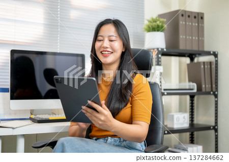 Asian woman freelancer office worker holding looking at tablet sitting on leather chair in company. Asian woman freelancer office worker holding looking at tablet sitting on leather chair in company. 137284662