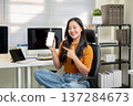 Pretty asian woman holding pointing at white screen smartphone sits on leather chair at home office. 137284673