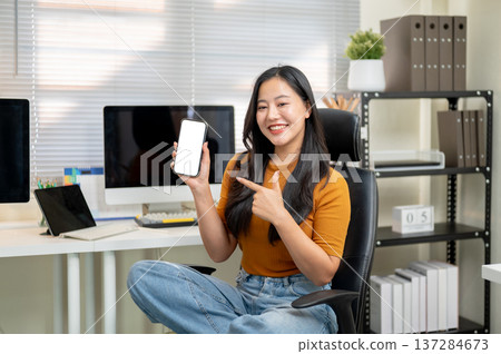 Pretty asian woman holding pointing at white screen smartphone sits on leather chair at home office. Pretty asian woman holding pointing at white screen smartphone sits on leather chair at home office. 137284673