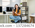 Pretty asian woman holding and using smartphone sitting folding legs on leather chair at home office 137284674