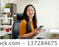 Smiling asian woman office worker holding smartphone as sitting on leather chair at home or company. 137284675