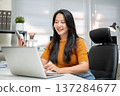 Asian woman freelancer or office worker holding phone looking at laptop as sitting at company table. 137284677
