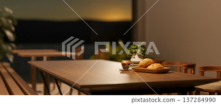 Bread and coffee with pot plant on wooden table in dim cafe dining room with sunrise outside window. Bread and coffee with pot plant on wooden table in dim cafe dining room with sunrise outside window. 137284990