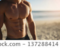 faceless athletic man front facing showing defined abs on sandy beach, ocean horizon softly blurred 137288414