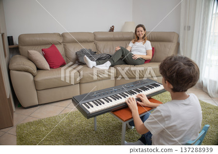 Son playing an electronic keyboard while his mother relaxes on the sofa, supporting his musical hobby and enjoying a moment of family bonding and learning in their cozy living room 137288939