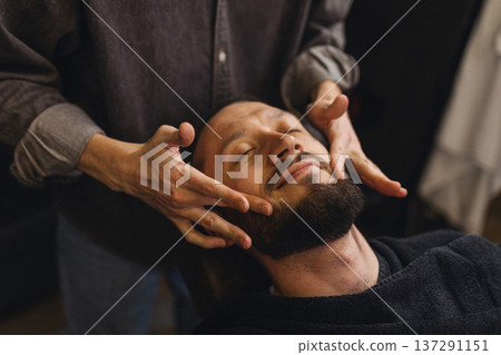 Barber massaging beard and neck of male client after shaving in barbershop. Relaxing grooming service, men self care, comfort and classic barber ritual concept. Barber massaging beard and neck of male client after shaving in barbershop. Relaxing grooming service, men self care, comfort and classic barber ritual concept. 137291151