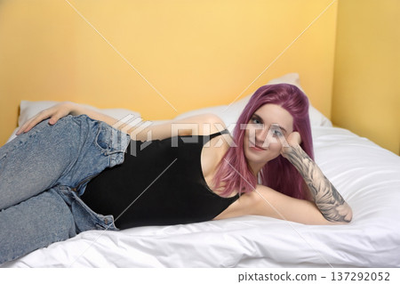 Smiling pregnant young girl with purple hair is lying on the bed. Smiling pregnant young girl with purple hair is lying on the bed. 137292052