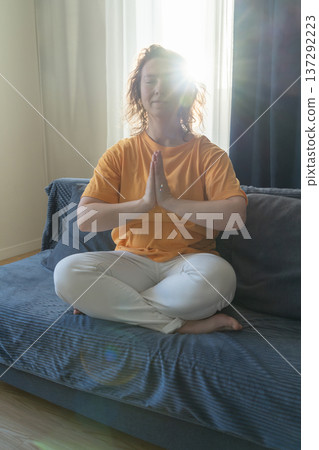 Woman sits in lotus position with her hands folded in prayer on sofa under bright ray of sunlight, practicing meditation and finding inner peace at home Woman sits in lotus position with her hands folded in prayer on sofa under bright ray of sunlight, practicing meditation and finding inner peace at home 137292223