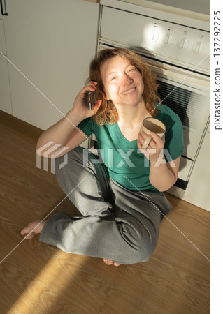 Young red-haired woman sits on the kitchen floor, smiling, talking on her smartphone, and drinking coffee Young red-haired woman sits on the kitchen floor, smiling, talking on her smartphone, and drinking coffee 137292225