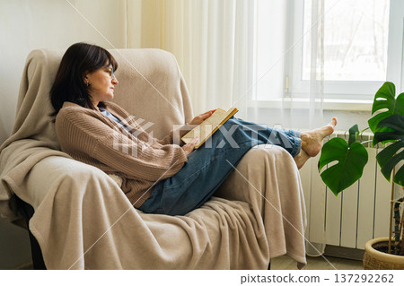 Woman in glasses relaxing in an armchair by the window, engrossed in a book, enjoying leisure time at home 137292262