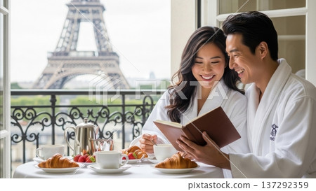 Asian couple in white bathrobes reading book together enjoying breakfast with croissants coffee and strawberries on Paris balcony with Eiffel Tower view 137292359