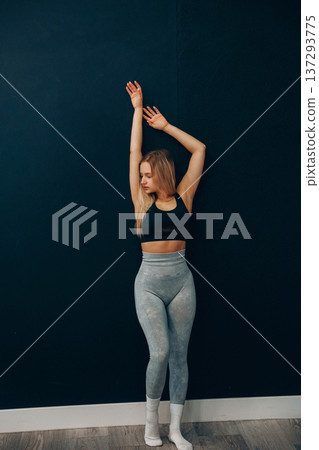 A young woman stretches by a wall wearing activewear, smiling while engaging in exercise for health and fitness. A young woman stretches by a wall wearing activewear, smiling while engaging in exercise for health and fitness. 137293775