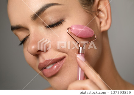 Woman using natural pink quartz face roller on grey background, closeup. 137293839
