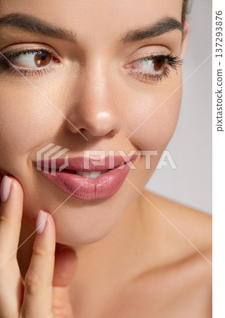 A woman smiles softly, showcasing her flawless skin and light pink lip color. Her hand gently touches her cheek, emphasizing her natural beauty and confidence. 137293876