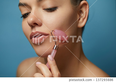 Woman using natural pink quartz face roller on blue background, closeup. Face massage.. Woman using natural pink quartz face roller on blue background, closeup. Face massage.. 137293886