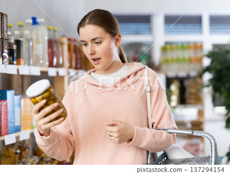 Girl customer in supermarket examines jar of pickled green olives, chooses food-stuff in grocery 137294154