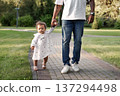 Father holding little daughter hand while child takes first steps on park path. Dad with baby outdoors 137294498