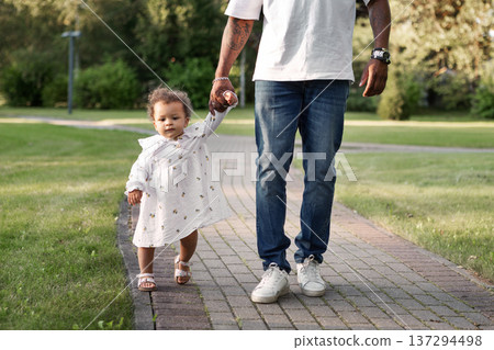 Father holding little daughter hand while child takes first steps on park path. Dad with baby outdoors 137294498