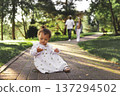 Curious little daughter sitting on park path and exploring nature while parents walk behind. Family lifestyle outdoors, childhood independence, care and safety. Small child at summer day in green park 137294502