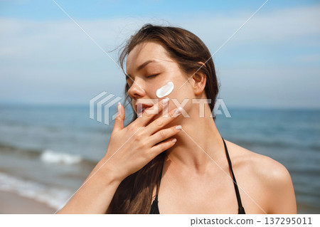 Attractive female applying sunscreen lotion on face with closed eyes at seaside, beauty skincare and sun protection concept with relaxed summer mood Attractive female applying sunscreen lotion on face with closed eyes at seaside, beauty skincare and sun protection concept with relaxed summer mood 137295011